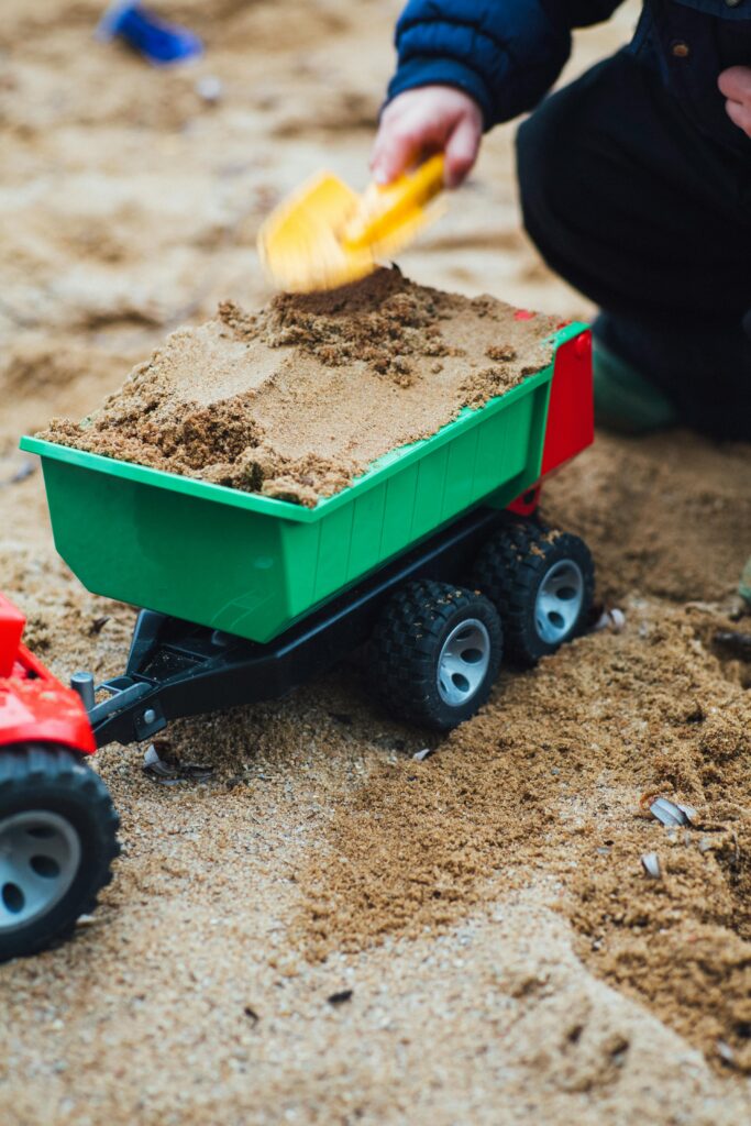 Young child using a shovel to fill a toy tractor with sand outdoors.