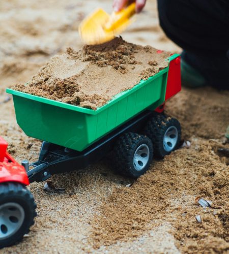Young child using a shovel to fill a toy tractor with sand outdoors.